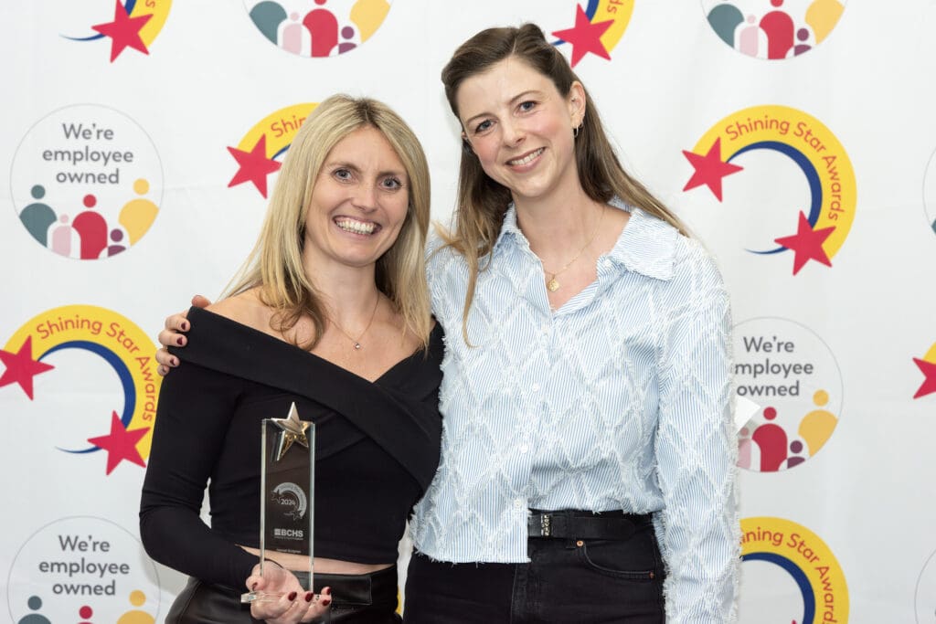 Woman smiling holding trophy next to another woman in front of a branded background