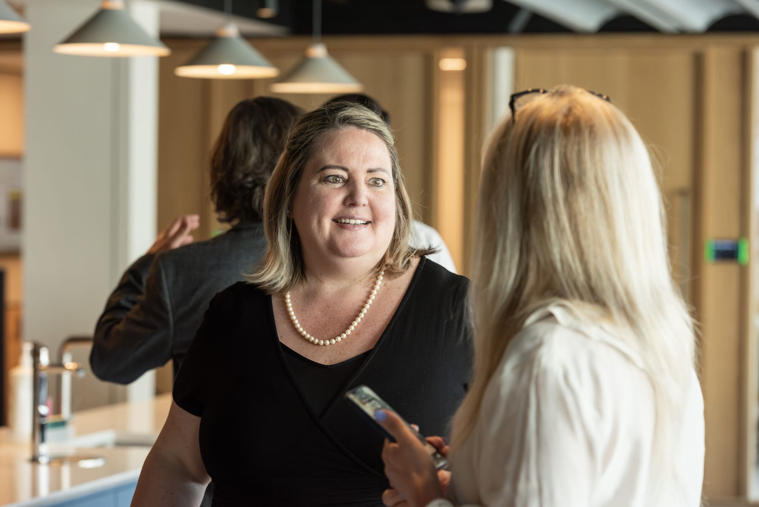 Two females in room of people talking to each other one facing the camera smiling